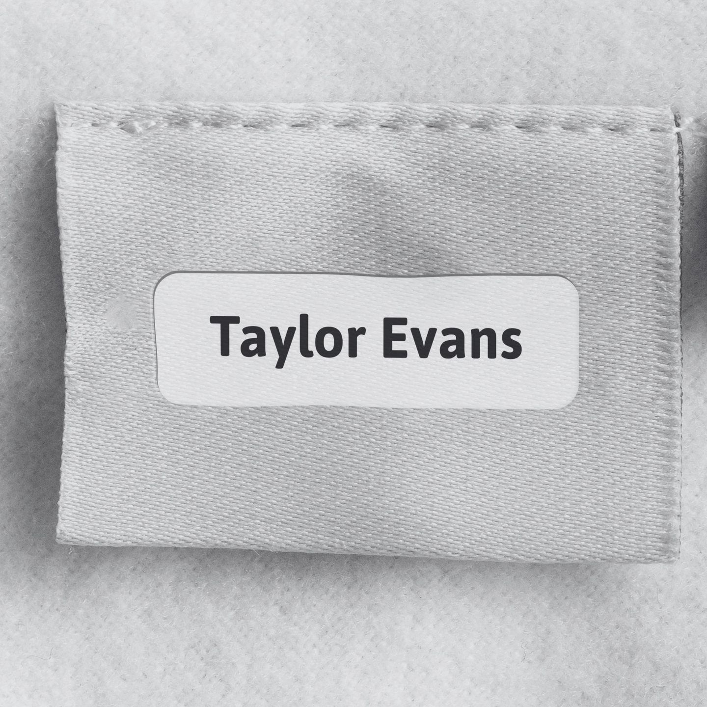 A close-up of a light gray fabric pouch or label holder with a white rounded rectangular patch reading 'Taylor Evans' in black text, photographed on a matching light gray textured background. personalised name tag labels